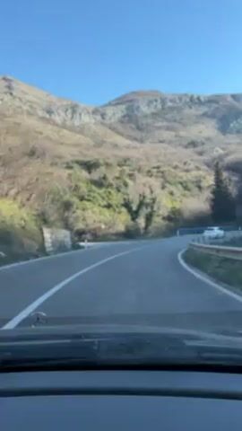 The video shows a drive on a paved road through a mountainous landscape under a clear, bright blue sky. The camera is positioned inside a car, looking out through the windshield. The car drives on a curved road with a white dividing line. The landscape features hills covered with sparse vegetation, and rock formations are visible on the right side. The road is flanked by blue metal guardrails. Other cars are seen driving in the opposite direction. The dashboard of the car is visible in the foreground, including the Mercedes-Benz emblem.  The general atmosphere suggests a calm, scenic drive.