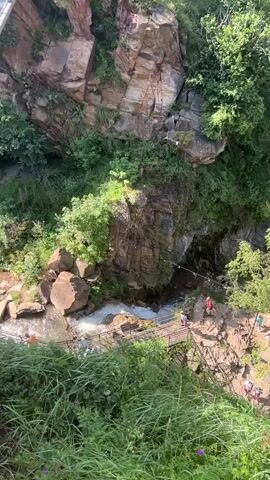 The video shows a high-angle view of a natural gorge with a river flowing through it. Several people are visible on a path and a wooden bridge constructed over the river. A person in a red shirt is standing on the bridge. The gorge is covered in lush green vegetation and rocky cliffs. In the background, houses are visible on a hillside. The overall scene depicts an outdoor recreational area, possibly a tourist attraction.