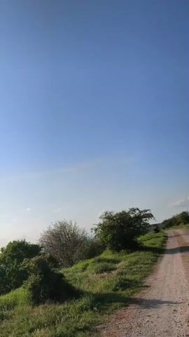 The video shows a landscape on a sunny day with a blue sky and a few clouds. The landscape features rolling green hills and fields, some of which are yellow. There are trees and bushes scattered throughout the scene, and in the distance, there are some buildings and a horizon line. The camera pans across the scene, revealing more of the landscape and the sky. The sun is visible in the sky, and there is a slight glare from it.