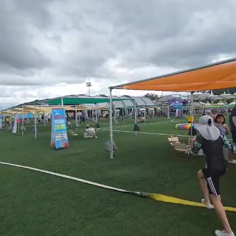 A drone shot pans across an outdoor market with numerous stalls set up on a green artificial turf field. The stalls are covered by canopies of various colors, including white and green, and orange. People are walking between the stalls, browsing or interacting. In the foreground, a person wearing a dark swimsuit and a wide-brimmed hat is walking away from the camera. Further back, a man is pushing a stroller with a child in it. The sky is cloudy.