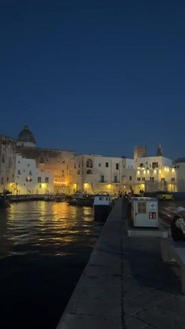 The video shows a waterfront at dusk or night. A body of dark water is in the foreground, reflecting lights from buildings on the opposite shore. These buildings are clustered together and appear to be made of white or light-colored stone, with windows illuminated by warm yellow light. Several boats are moored in the water or moving slowly. A domed building, possibly a church or cathedral, is visible among the structures. The sky above is a deep, clear blue. In the immediate foreground, a stone walkway or pier extends into the frame, with a gray surface. A few dark, indistinct figures are visible seated on a bench in the lower right corner, partially obscured. There is also a white utility box with some signage on the pier. The overall atmosphere is peaceful and serene, illuminated by artificial lights.