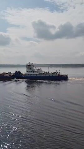 The video shows a long, blue and white tugboat with a large barge attached to it moving across a body of water. The barge appears to be carrying some sort of material, possibly lumber or construction supplies. The water is dark and rippled, and the sky above is partly cloudy.  The tugboat is creating a wake as it moves, suggesting it is moving at a moderate speed.  Trees are visible on the shoreline in the distance.