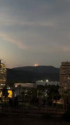 The video shows a sunset over a hilly landscape. The sun is partially obscured by a mountain peak, creating a ring of light. In the foreground, there are buildings and palm trees. The sky is cloudy and gray, with some lighter clouds visible. The overall mood is peaceful and serene.