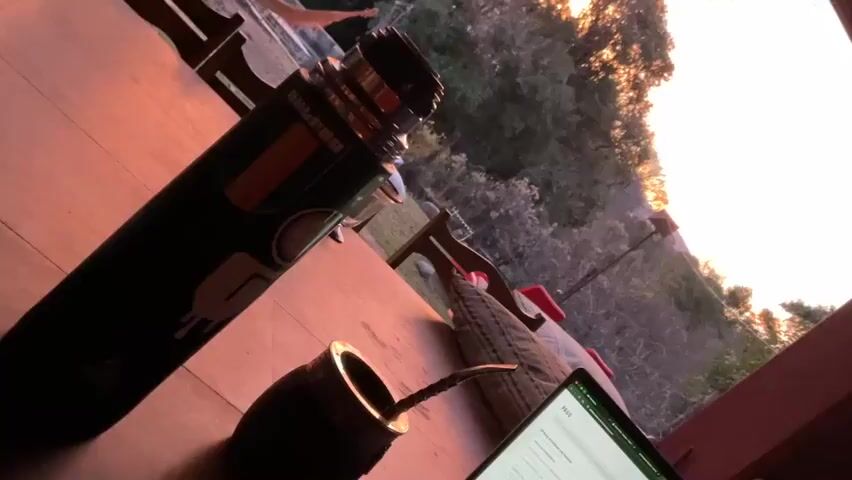 The video is a close-up shot of a mate gourd and a thermos on a wooden table. In the background, a hammock hangs between trees, and a laptop is open on the table. The sun is setting, casting a warm glow over the scene. The video is filmed at eye level.