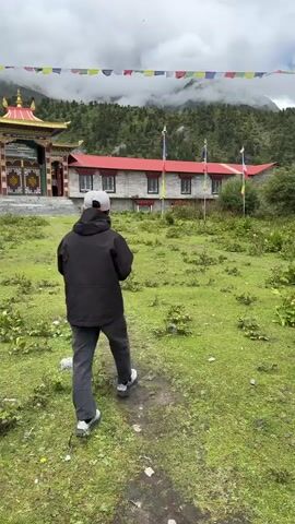 A person in a black jacket and gray pants with a gray baseball cap walks away from the camera on a dirt path through a grassy field. Behind them is a building complex with red roofs and stone walls. A large, ornate entrance structure with a gold roof is on the left side of the complex, with large double doors. Prayer flags are strung across the top of the scene. Mountains covered in green trees and shrouded in clouds are in the background.