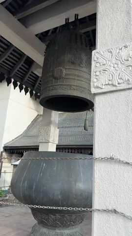 The video begins with a close-up of a large bronze bell hanging from a wooden structure. The bell is ornate and appears old. Next to it is a large dark bowl, possibly for collecting offerings. The camera then pans up and to the right, revealing a white-painted wall with decorative carvings and chains. The scene transitions to an outdoor view of white-walled structures with arched openings, adorned with various Buddhist flags. Palm trees and lush green foliage are visible in the background. The sky is partly cloudy with patches of blue. The camera continues to pan, showing more of the white structures, flags, and the surrounding natural landscape, including a hill covered in trees. The video ends with a wider shot of the environment. There are no people clearly visible in the video. The overall setting appears to be a religious or historical site, likely in a tropical region.