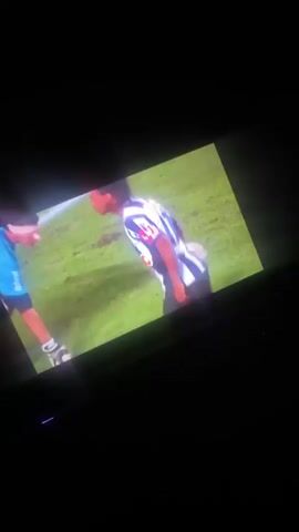 A video is being played on a screen in a dimly lit room. The screen displays two distinct scenes, both featuring a dark-skinned male, likely the famous Brazilian football player Ronaldinho. Bright glare from an external light source reflects off the television screen throughout the clip, partially obscuring the view.

The first scene (0:00-0:03) shows the player on a football pitch. He is wearing a blue and white striped football jersey with "MRV" and "BMG" sponsor logos prominently displayed. His dark hair is styled in dreadlocks or long braids, pulled back. He stands on a green grass field with white lines, and another blurry figure is partially visible to his left. He appears to be looking towards the left, perhaps surveying the field or an ongoing play.

The second scene (0:03-0:05) transitions to the same individual, now seated at a press conference table. He is wearing a dark, short-sleeved shirt with green and red geometric patterns and a green beanie hat. He leans forward, speaking into a microphone. The background is a step-and-repeat banner filled with various sponsor logos, including "PUMA," "BRAMHA," "BMG," "TIM," "MRV," "GATORADE," and "VGALO HD."

French audio is audible, with a male voice stating, "Qu'est-ce que vous comptez faire?" (What are you going to do?) and "Mon frère travaille là-dessus en ce moment." (My brother works on it at the moment.). The viewing environment suggests the video is being watched during nighttime or in a dark interior.