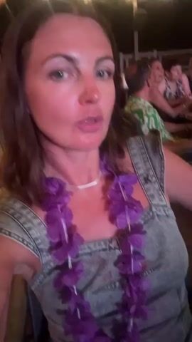 A woman with brown hair, wearing a denim overalls and a purple lei, is speaking to the camera. Behind her, several people are seated at tables in what appears to be an outdoor restaurant. The environment is dimly lit with string lights overhead. The woman is animated and appears to be enjoying herself. The scene captures a moment of celebration or a social gathering.