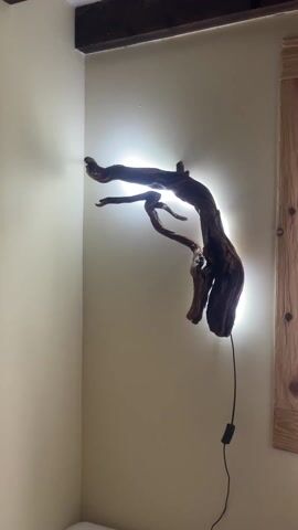 The camera slowly pans from a wall-mounted lamp made of a piece of driftwood to a window. The window is framed with wooden trim and covered with white sheer curtains.  A small table with a lampshade on it is visible in front of the window. Above, there are dark wooden beams on the ceiling. The wall is off-white. The bed is visible at the bottom of the shot.