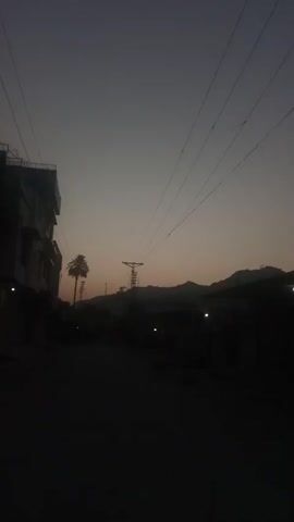 The video captures a street scene at dusk or dawn, with the sky displaying a gradient of muted colors. Power lines are prominently visible, stretching across the frame. On the left, a multi-story building is partially visible, its architecture suggesting a residential or commercial space. In the mid-ground, a lone palm tree stands out against the dusky sky, and a tall electrical pylon is positioned further down the road. The street itself appears to be empty, with the dim light casting long shadows and obscuring finer details of the surrounding buildings and terrain. The overall atmosphere is one of quietude and fading light.