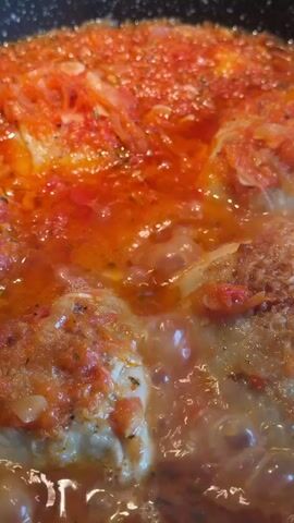 This is a close-up, overhead shot of food cooking in a pan. The food appears to be chicken breasts covered in a thick, red sauce made with tomatoes and possibly onions. The sauce is bubbling, indicating it is simmering or boiling. Some pieces of chicken are visible beneath the sauce. The pan is black. The lighting suggests it is an indoor setting, likely a kitchen.