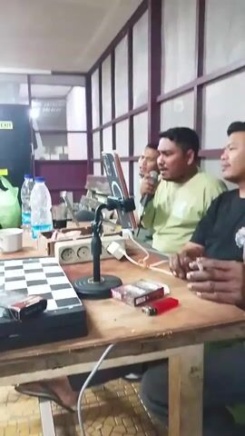 Three men are seated at a wooden table. The man in the center is holding a microphone to his mouth and appears to be singing or speaking. He is wearing a light green t-shirt. To his left, another man is partially visible, looking towards the center. To his right, a man wearing a black t-shirt is looking towards the center and holding something near his mouth, possibly a cigarette. On the table, there are several items including a chessboard, two packs of cigarettes, a lighter, a white mug, and two bottles of water. A mobile phone is propped up on a stand in front of the center man. In the background, there is a wall with vertical partitions, and an "EXIT" sign is visible on a door. The lighting appears to be artificial.