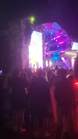 This is a nighttime outdoor concert scene. A large stage with a bright video screen is set up. The screen displays a person, possibly a performer, in a yellow outfit. The stage is illuminated by colorful lights, including purple and blue. A crowd of people are gathered in front of the stage, facing the performance. Many people are silhouetted against the bright lights. To the right of the frame, a dark vehicle, possibly a bus, is parked, its red taillights illuminated and casting a red glow on the ground. The overall atmosphere appears festive and energetic.