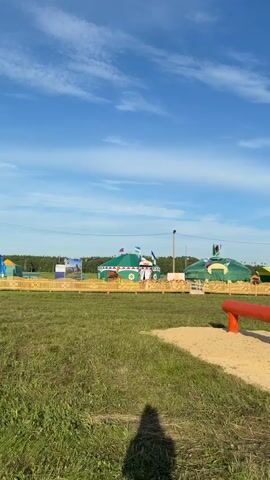The video shows an outdoor scene with a clear blue sky and scattered clouds. In the foreground, there is a patch of sand with a red horizontal beam, possibly a practice obstacle, and a shadow of a person is cast on the grass. In the midground, there is a grassy field with several large, colorful tents or yurts, some with flags. A wooden fence encloses an area in front of the tents. The background shows trees and a long fence line. The overall impression is of a festival or event grounds.