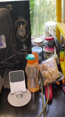 The video shows the interior of a dismantled television. The camera pans across the various electronic components, including circuit boards, wires, a speaker, and the cathode ray tube. The objects in the foreground are a cell phone stand, a water bottle, and a drill. The background appears to be an outdoor setting with greenery and some containers. The scene is cluttered and dusty, suggesting a workspace or storage area. There are no people visible in the video.
