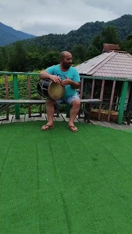 A man sits on a wooden bench in an outdoor setting and plays a drum. The man is bald, wearing a teal t-shirt and blue shorts. He is holding a black and tan drum and appears to be hitting it with his hands. The bench is on a wooden deck with a green, carpet-like surface in front of it. In the background, there is a wooden railing, a small building with a brown roof, and a mountain range covered in trees. The sky is overcast.
