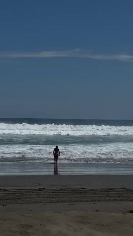 A long shot captures a person wading into the ocean from a sandy beach on a sunny day. The sky is clear blue with a few faint clouds. White waves break along the shoreline, extending into the distance. The foreground shows dry sand with horizontal tire-like tracks, transitioning to wet sand where the person is. The individual, dressed in a dark swimsuit, walks steadily forward, facing away from the camera, into the incoming waves that reach their waist.