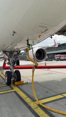 The video shows the front section under an airplane. The plane's landing gear is visible, with two wheels chocked by yellow blocks. A thick, yellow cable is connected to a point near the engine. The aircraft is white with a red and white tailfin visible in the background, along with other airport vehicles and equipment. The plane sits on a gray concrete tarmac marked with yellow lines.