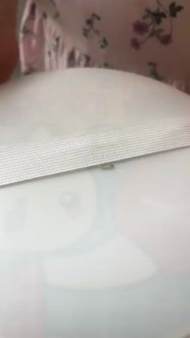 A hand with decorated fingernails is shown interacting with a small, light-colored object on a white surface. The object appears to be a worm or larva. The fingernails have colorful designs, including what looks like apple motifs and floral patterns. A white elastic band is stretched horizontally across the frame, above the object. The white surface has a faint cartoonish pattern of a face with large eyes. The background shows a person wearing a light pink dress with a floral pattern. The hand is seen touching the object with a fingernail. The lighting is bright and even.