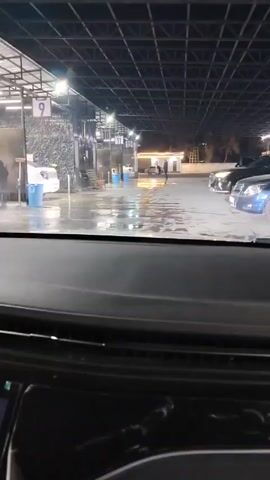 The video is shot from the interior of a vehicle parked in an open-air car wash. The car wash is covered by a metal grid roof with lights. A person is washing a white sedan with a hose in one of the car wash bays. There are other cars parked in the vicinity. The dashboard of the car the video is taken from is visible. A digital clock on the car's display reads 21:54.