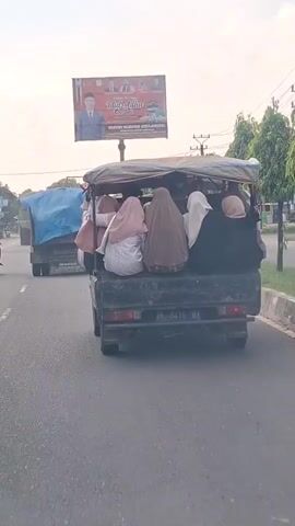 A rear view of a pickup truck driving on a road. The truck bed is covered with a tarp and filled with people, most of whom are wearing hijabs. A billboard is visible in the background. The truck is in motion, and the camera appears to be following it. The scene is outdoors during the day.
