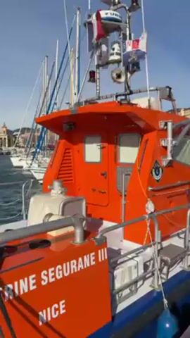The video shows the view from the deck of a bright orange and white boat as it leaves a harbor in Nice, France. The boat is marked "E Segurane III" and "Nice". A small cabin is on the back of the boat with a radar and a few small flags on top. Several masts of sailboats are visible nearby, docked in the harbor. In the background is the cityscape of Nice, including a large clock tower, and green hills on the edges of the city. The water is a deep blue, with some wakes visible.