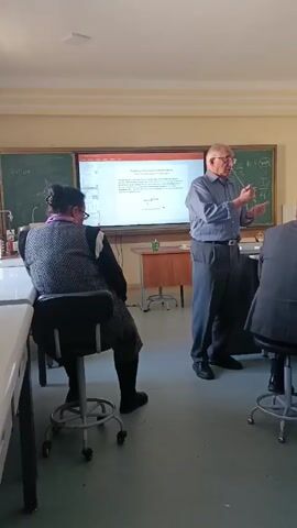 In a bright, sunlit classroom, an elderly man in a blue shirt and gray slacks stands presenting in front of three seated individuals. The presenter stands in front of an interactive whiteboard displaying a diagram and text. A chalkboard with handwritten equations is behind the whiteboard. The students are seated on rolling stools. The room has rows of white desks. The overall tone is academic and professional.