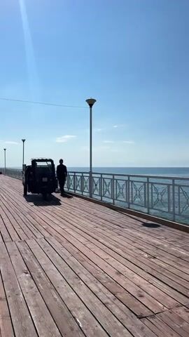 A person is walking on a wooden pier next to a black three-wheeled vehicle. The pier has a white railing along the water's edge. A bright sun is in the sky, and the water is blue. There are streetlights along the pier. The video appears to be taken on a sunny day.