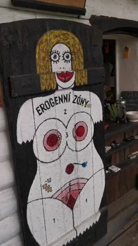 The video shows a wooden board painted to resemble a woman's body, used as a dartboard. The board is predominantly white with black outlines, depicting a cartoonish woman with blonde hair, blue eyes, and red lips. The top section of the board has the words "EROGENNÍ ZÓNY" (Erogenous Zones) written in black. Various parts of the body are numbered with different point values, ranging from negative numbers to positive numbers like 1, 2, 3, 5, and 10. A dart with a blue flight is seen stuck near the center of the abdomen, while another dart with a red flight is stuck in the groin area. The background shows parts of a building, possibly a bar or restaurant, with shelves, bottles, and other items visible. Laughter and excitement can be heard as the darts are thrown.