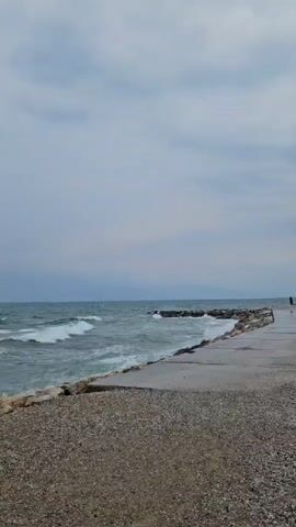 The video shows a rough sea with waves crashing against a rocky shore. The sky is overcast and the water is a murky green. The camera is positioned on the shore, looking out at the sea. There are no people or animals visible in the video. The scene appears to be a natural landscape, possibly a coastal area. The overall mood of the video is somber and dramatic, due to the rough sea and overcast sky.