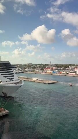 The video shows a large cruise ship, the MSC Seaside, docked in a harbor. The ship is white with many decks and large windows. It is tied to a small floating platform with several people on it. The water is a clear turquoise color. In the background, there is a town with buildings of various colors. The sky is blue with scattered white clouds. The scene appears to be taken on a sunny day.