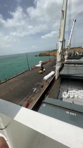 The video shows a cargo ship being loaded at a port. Two cranes are positioned over the ship's open cargo hold, which is filled with white bagged goods. A yellow forklift is visible on the dock, near stacks of the same white bagged goods. The water is a vibrant turquoise, and the sky is partly cloudy. In the background, a rocky coastline with some industrial structures is visible. A person is seen standing on the dock. The ship has the marking "19.00T" visible on its side.