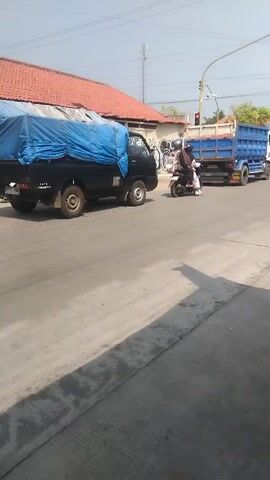 A black pickup truck covered with a blue tarp drives down a paved road. A blue dump truck follows behind it. A person on a motorcycle drives past the pickup truck. A building with a red tiled roof is visible behind the pickup truck. A street light is visible in the background. A man on a motorcycle carrying several large water jugs drives down the road. A construction site with an excavator and construction materials is visible in the background.