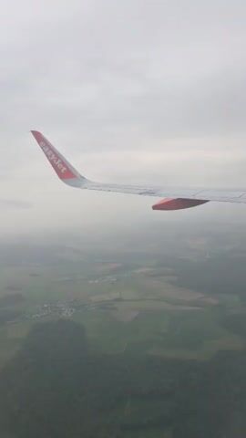 The video shows a view from an airplane window. An EasyJet plane wing is visible on the right side of the frame. Below, there is a landscape of green fields, some wooded areas, and a small town. The sky is overcast with a grey cloud cover. A large bridge is visible in the distance.