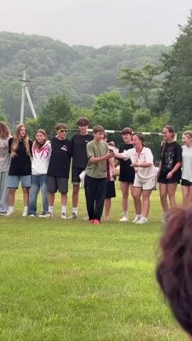 A group of approximately ten young people, appearing to be teenagers, stand in a grassy field. The foreground shows a blurred, out-of-focus dark object, possibly a person's back or shoulder. In the background, there is a line of trees and a hill covered in vegetation. The sky is overcast.

The individuals are arranged in a line, facing towards the viewer and slightly to the left. The person in the center, a male wearing a green t-shirt and black pants, is holding a smartphone and appears to be speaking or performing. His mouth is open, and his right arm is extended. Other individuals in the group are looking at him, some with smiles, and some are clapping or gesturing. The person to the right of the central figure, a female wearing a white t-shirt and shorts, is clapping enthusiastically. A person on the far left, a female with long blonde hair, is looking forward. A male on the far right is also looking forward.

Clothing is casual, including t-shirts, shorts, and jeans. Some individuals are wearing sneakers. The overall atmosphere appears to be informal and possibly an outdoor event or gathering. There is a faint suggestion of a soccer goal in the far background. The lighting is diffused due to the overcast sky.