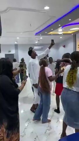 The video shows a group of people dancing and enjoying themselves in a large, well-lit hall. Some people are taking videos with their phones, while others are clapping and cheering. The atmosphere appears celebratory. The hall has a modern design, with a decorated stage area in the background. Many people are standing and watching, seemingly enjoying the music and dancing. Chairs are lined up in rows, indicating a possible event or gathering. The people are diverse in age and appearance, dressed in casual and semi-formal attire.