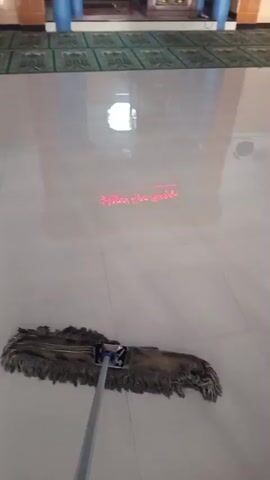 A first-person perspective video shows a person cleaning a tiled floor with a dusty mop. In the background, rows of green prayer mats are visible along a wall with pillars and ornate wall decorations. A red laser line projects onto the floor, suggesting a measurement or alignment tool. The floor is highly reflective, with a bright light source causing glare. The action is limited to the movement of the mop across the clean, tiled surface.