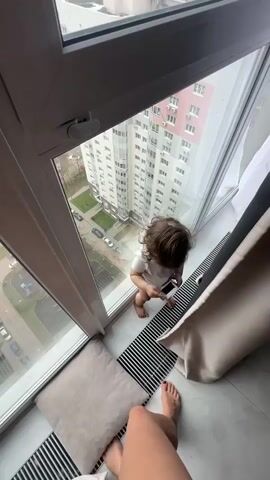 A high-angle shot shows a baby standing in a window frame looking out over a city. The baby is wearing a white shirt and has curly brown hair. The window frame is white, and there is a large window pane to the left and above the baby. Outside the window is a view of a city with tall apartment buildings. A woman, presumably the baby's mother, is adjusting the curtains on the right side of the frame. Her feet are visible in the lower part of the frame. Another toddler appears and looks out the window along with the baby. The baby laughs.