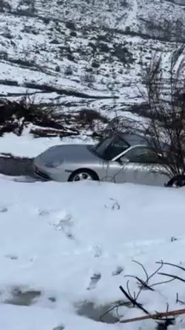 A silver Porsche 911 is parked on a snowy road. A blue and white Renault Captur is parked behind it. The road is on a hill, and the surrounding area is covered in snow. There are some bushes and trees visible in the background. The video is shaky. The camera pans up and zooms out, showing the cars parked on the side of the snowy road.