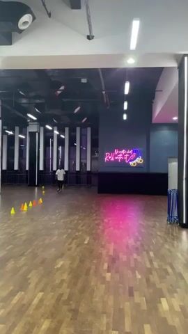 A person wearing a white t-shirt and dark pants is roller skating around a series of orange and yellow cones on a wooden floor in a large indoor space. The space has a dark ceiling with fluorescent lights and some decorative elements hanging from it. A neon sign that says "No matter what, Roll with it" with a graphic of roller skates is visible on a dark blue wall in the background. To the right, there are some blue rolling racks. The lighting is dim in some areas and brightly lit in others, with a pinkish-purple glow on the floor in the foreground. The person is moving from left to right in the frame, navigating through the cones.