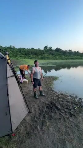 The video opens with a man in a grey tank top, black shorts, and green boots standing on muddy ground beside a light-colored tent. He has tattoos on his left arm and is looking towards a body of water on his right. Another man in an orange shirt is visible behind the first man, kneeling near the tent. A fishing rod is upright near the standing man. The camera slowly pans right, revealing a calm body of water reflecting the sky and distant treeline. Two fishing rods are seen extended from the muddy bank into the water. The sky shows a soft blue at the top, transitioning to a pale pink and orange hue at the horizon.
