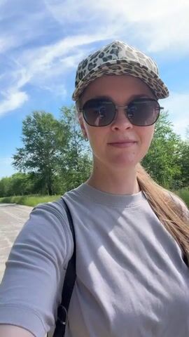 A woman with long brown hair, wearing sunglasses and a leopard-print cap, films herself walking on a paved path outdoors. She holds a stick in her right hand. A young boy rides a green bicycle with training wheels on the same path, slightly ahead of her and to her right. The boy is wearing a striped shirt, blue shorts, and a patterned bucket hat. The environment appears to be a park or recreational area with trees and grass on either side of the path. The sky is blue with some clouds. The woman is speaking, but her words are inaudible.