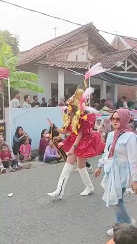 A street parade is underway. In the center, a performer in a bright red costume with elaborate gold and pink feathered headdress and shoulder pieces dances. They wear white boots. To the right, a woman in a light blue shirt and a pink hijab watches. To the left, a young child in a pink outfit is visible. Various people are gathered on the sides of the street watching the parade, some sitting, others standing. Behind the performer, a pink tent with signage is set up, likely for a vendor. In the background, there are houses and trees. The sky appears overcast with hints of sunset lighting.