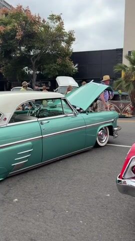 The video shows an outdoor event featuring classic cars. In the foreground, a red convertible is parked, its trunk open revealing a spare tire and other items. The car has a California license plate that reads Z 96 721 SAN DIEGO. To the left of the red car is a teal-colored convertible with its hood up. Further in the background, other classic cars and people are visible. A building with signs reading "ESCAPE ROOM" and "PHO GRIL" is behind the cars. The overall atmosphere suggests a car show or gathering.