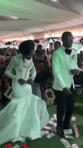 A bride and groom dance at their wedding reception, as guests throw money at their feet. The bride wears a long white mermaid-style gown with sheer lace sleeves and a high neck. The groom wears a light-colored tuxedo with a black bow tie. They are dancing on a green carpet covered with scattered paper money. The reception appears to be held in a tent or large room with a low, white ceiling. Tables and chairs are visible in the background, as well as other wedding guests. The general ambiance appears festive.