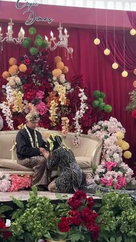 A man and a woman are seated on a couch during a wedding ceremony. The man is wearing a black top, patterned pants, and a headdress. The woman is wearing a dark, ornate dress with a head covering and is kneeling in front of the man, holding his hands. The background features a red curtain, elaborate floral arrangements in shades of red, pink, yellow, and green, and decorative lights. The setting appears to be an indoor event space decorated for a wedding.