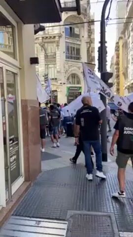 The video shows a group of people marching along a sidewalk in a city. The people are carrying flags and at least one person has a white umbrella. Some of the people are wearing black shirts. The buildings in the background are tall and ornate. The street looks wet.