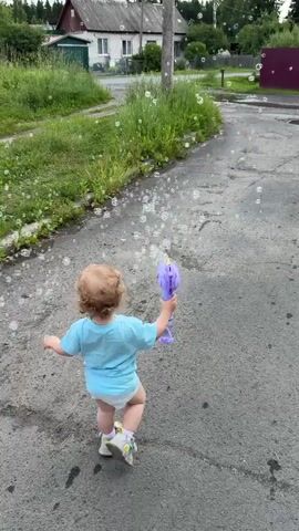A toddler with blonde curly hair, wearing a light blue t-shirt and a diaper, is walking on an asphalt path. The toddler is holding a purple bubble wand in their right hand, which is producing numerous soap bubbles. The toddler is facing away from the camera, looking towards the right side of the frame. In the background, there is a grassy area with some small white flowers, a wooden fence, a white house with a red roof, and a green shed. A gray utility pole stands to the right of the toddler. The sky appears overcast. The overall scene suggests a child playing outdoors in a rural or suburban setting.