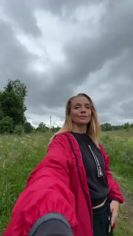 A woman with blonde hair, wearing a red jacket over a black sweatshirt, stands in a grassy field under a cloudy sky. She is holding a phone or camera in her right hand, appearing to film a selfie video. The field is overgrown with green grass and scattered white flowers. A dirt path runs through the field, leading into the distance. In the background, there are trees and a utility pole. The woman looks towards the camera and smiles. The video is shot from a low angle, looking up at the woman and the sky.