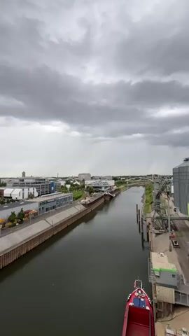 The video shows an aerial view of a canal or river with buildings lining its banks. The sky is overcast with dark, dramatic clouds. On the left side of the waterway, there are modern buildings with flat roofs, and a street with trees and parked cars. The canal itself is dark and appears calm, with a red and white boat docked on the right side. Further down the canal, there is a long, flat-bottomed barge. The cityscape in the background features a mix of modern and older architecture, including what appears to be a church with a prominent dome. Industrial cranes are also visible along the waterfront. The overall atmosphere is somewhat somber due to the cloudy weather.